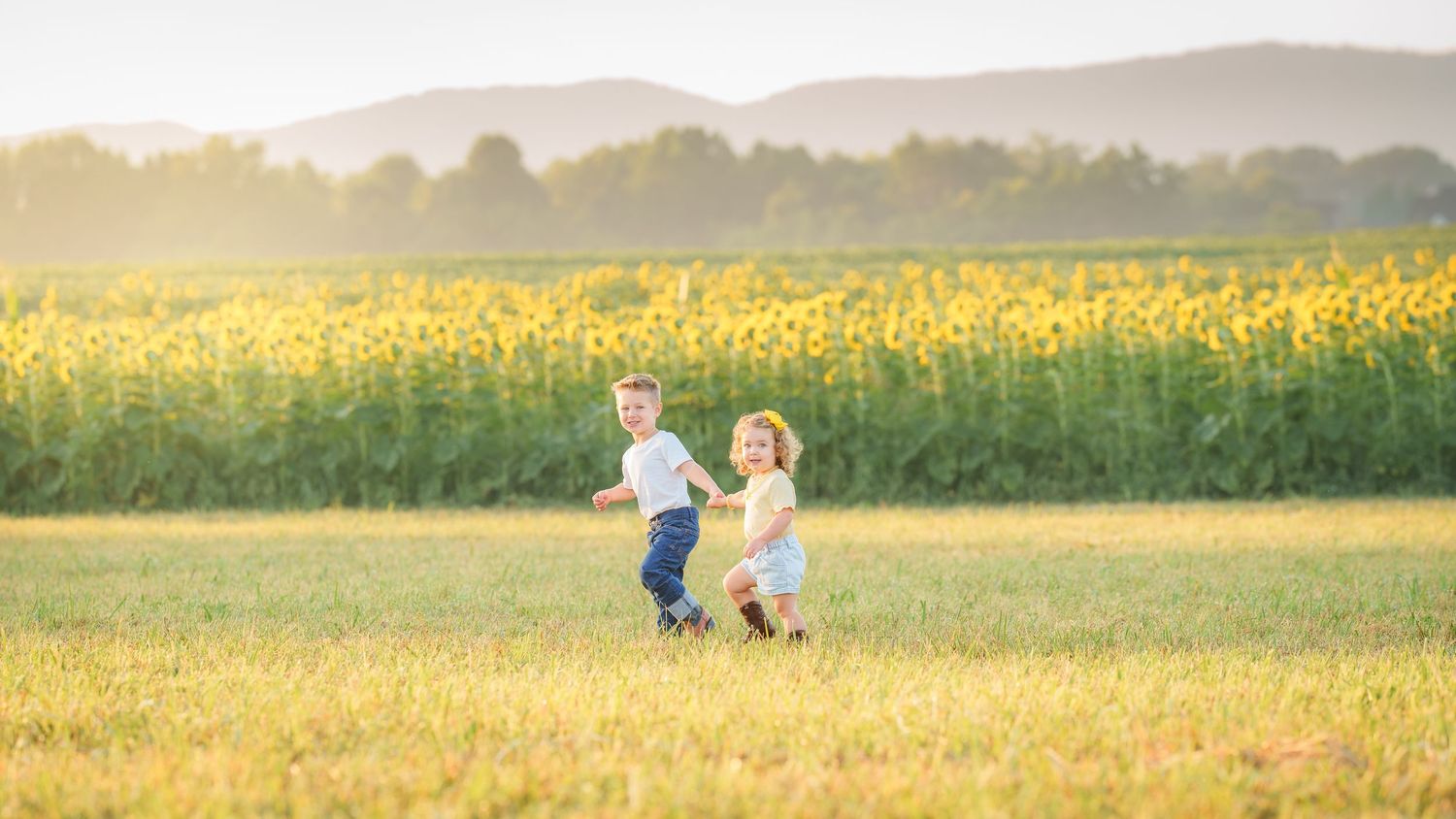 Two happy children running through a sunflower field on a sunny day with mountains in the background.