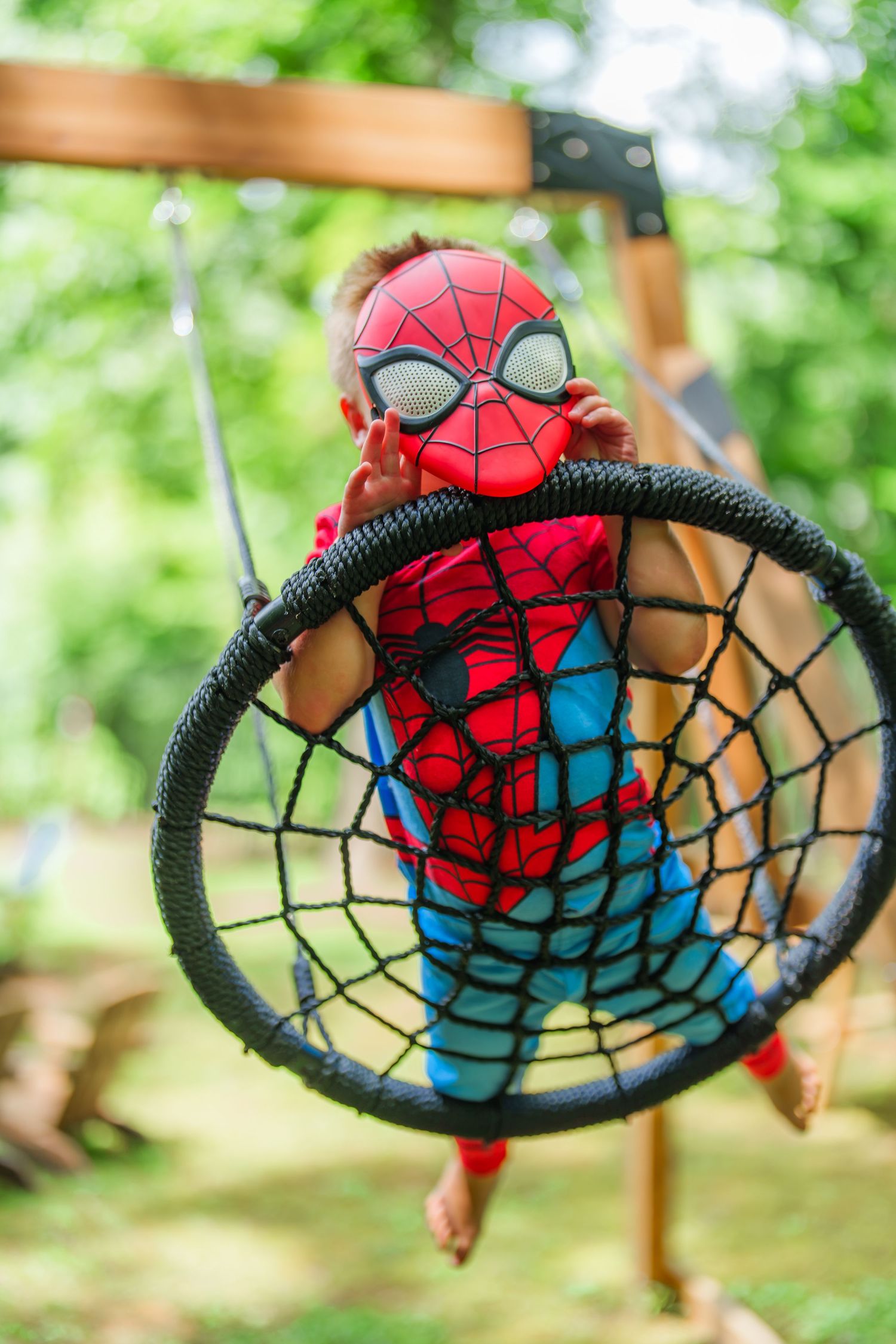 Child in Spiderman costume sitting in a spider web rope swing outdoors in a green backyard.