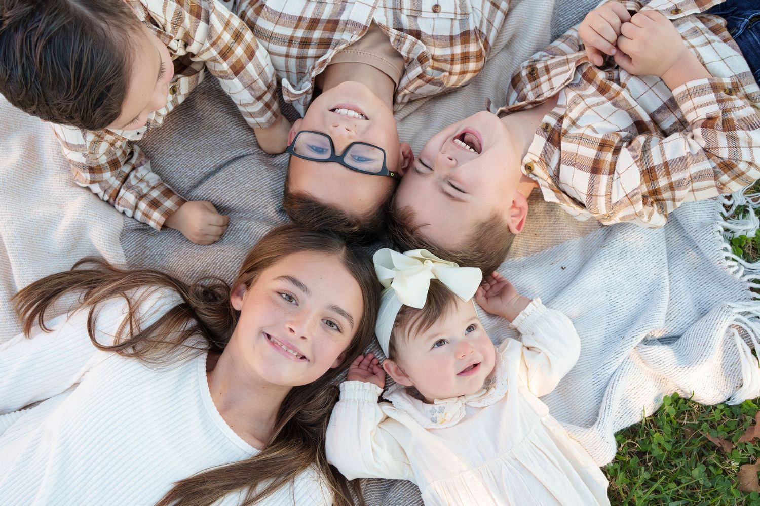 Five children lying in a circle on a blanket outdoors smiling up at camera.