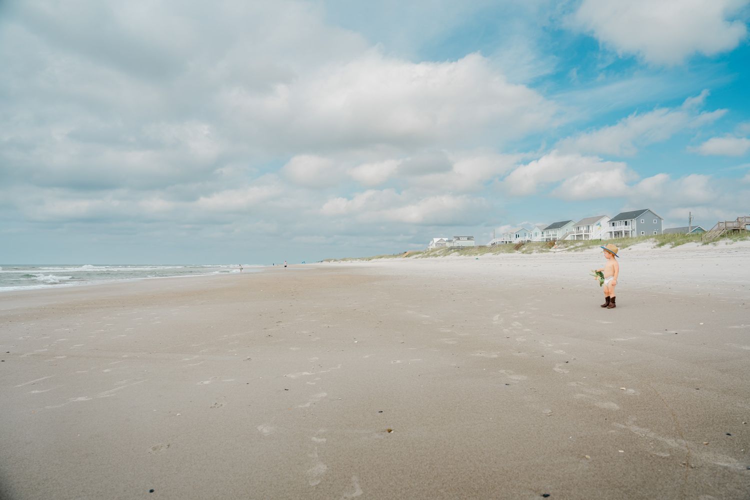 Toddler in a dress standing alone on a wide sandy beach under a cloudy sky with houses in background.