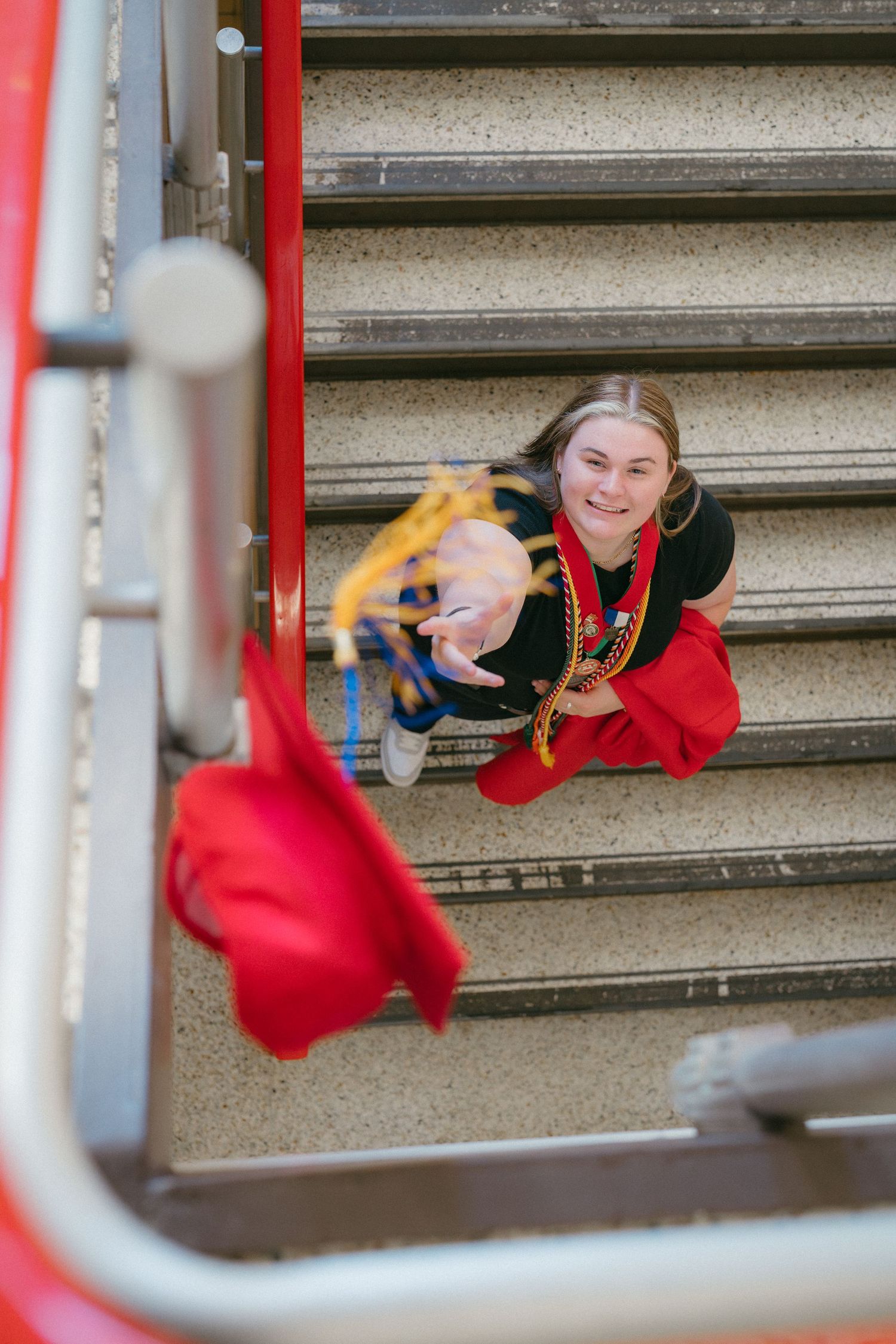 Overhead view of a smiling graduate in red gown sitting on outdoor stairs pointing upward.