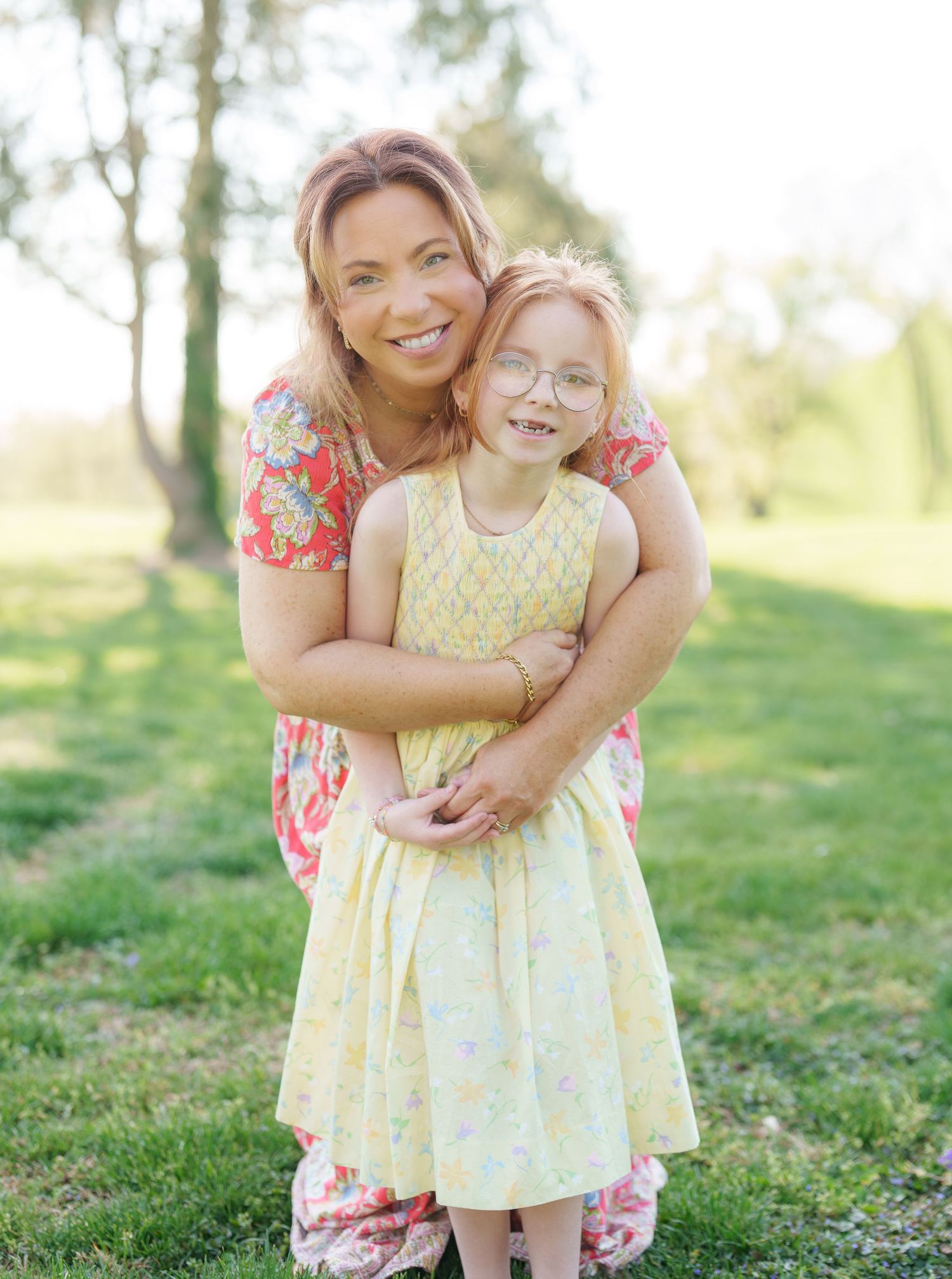 Smiling mother hugging young daughter wearing yellow dress and glasses in sunny green park.