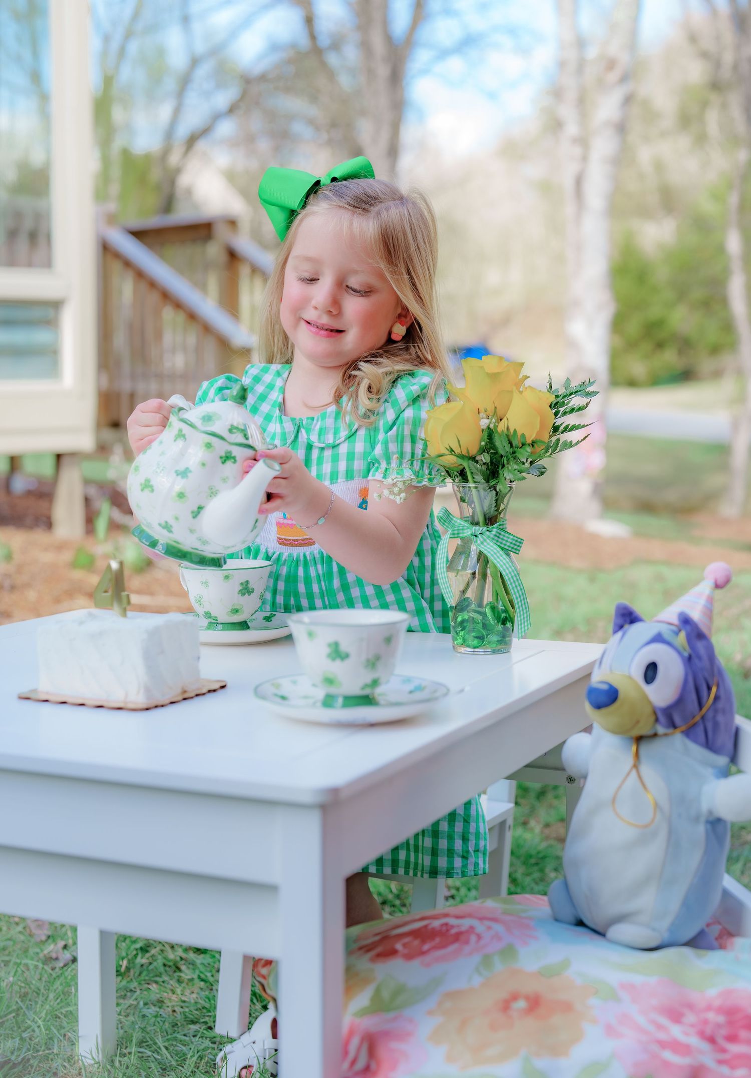 Young girl in green gingham dress pouring tea at a white table with yellow flowers and stuffed animal outdoors.