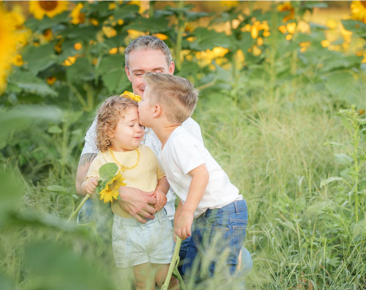Father kneeling in sunflower field with two young children hugging, surrounded by bright yellow sunflowers and green grass.