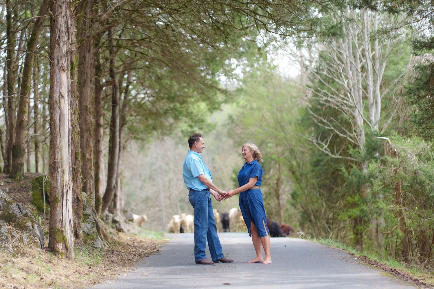 Couple holding hands on a tree-lined country road, dressed in blue, sharing a romantic outdoor moment.