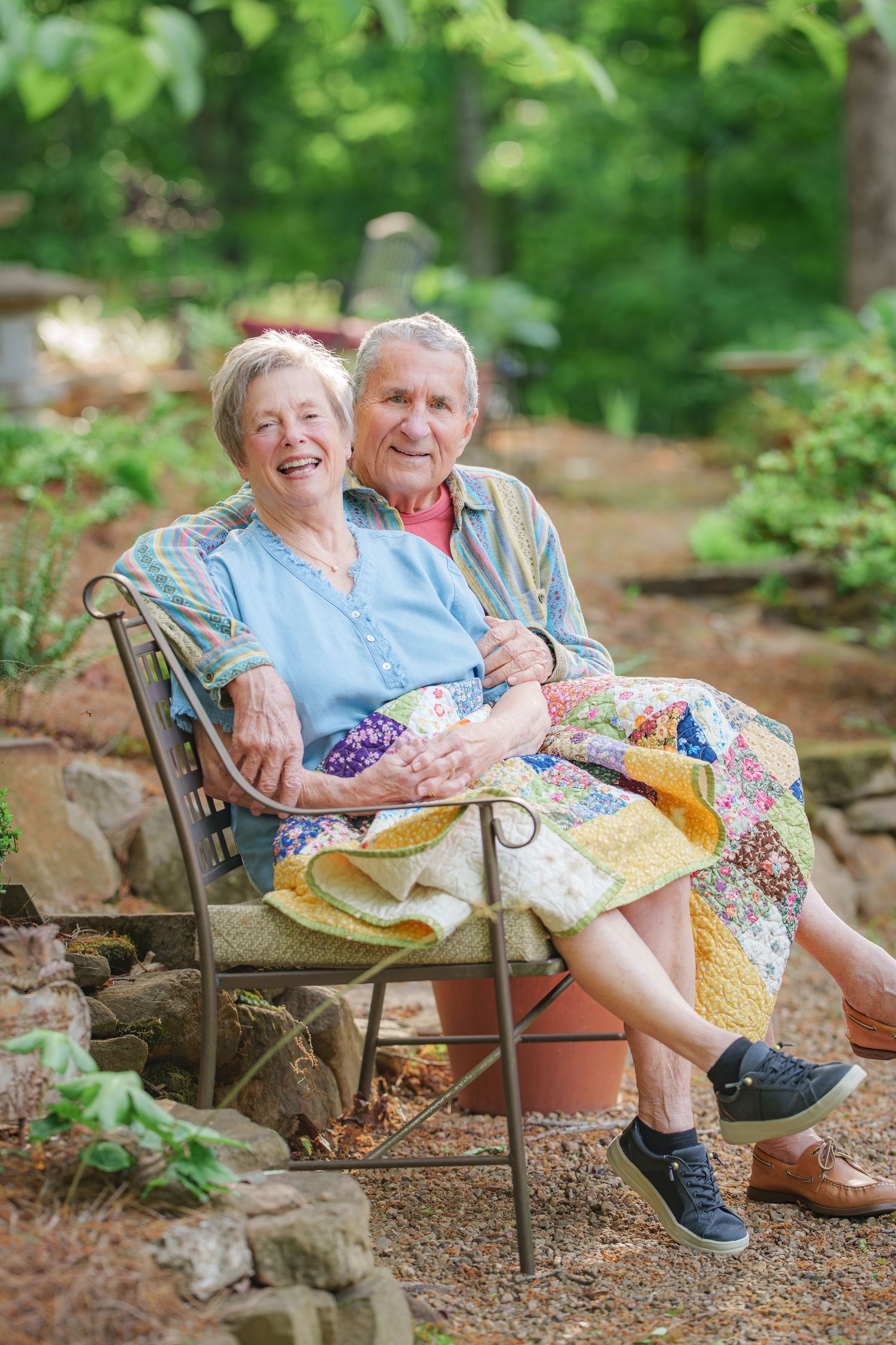 Smiling elderly couple sitting together on a bench with a colorful quilt in a garden.