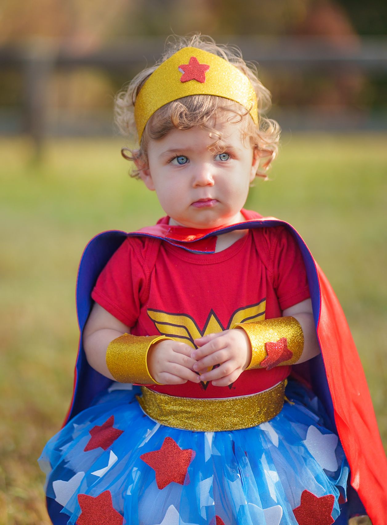 A cute toddler girl dressed in a Wonder Woman costume with cape and gold tiara poses outdoors in fall.