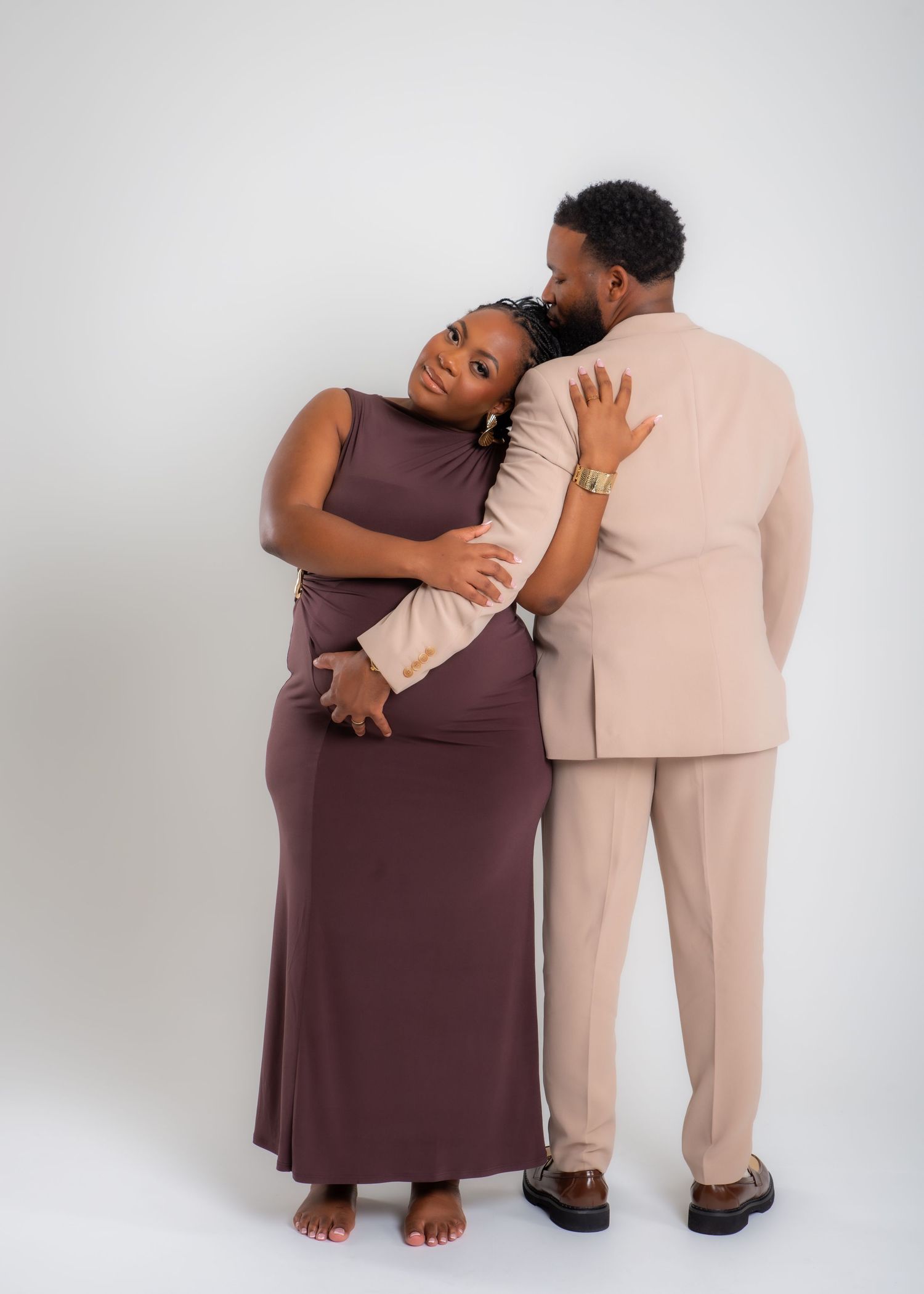 Two people wearing brown and beige outfits embrace in a tender moment against a white background.