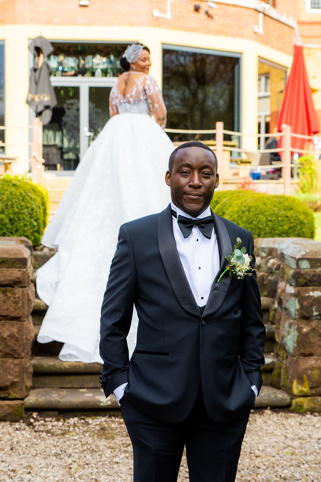 A groom in classic black tuxedo poses outdoors against a brick building backdrop.