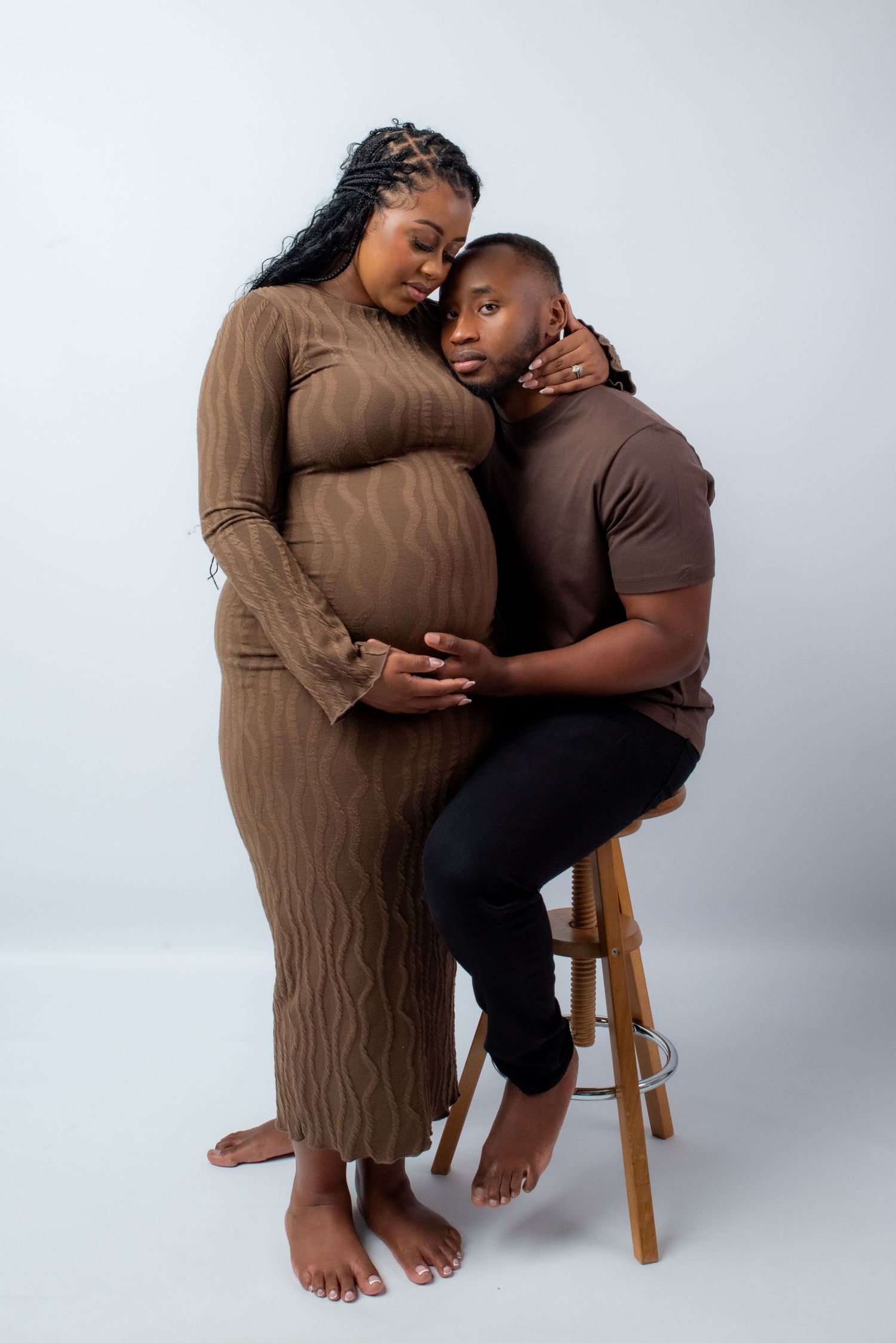 A tender maternity photoshoot showing an intimate moment between a couple in brown and black outfits against a light backdrop.