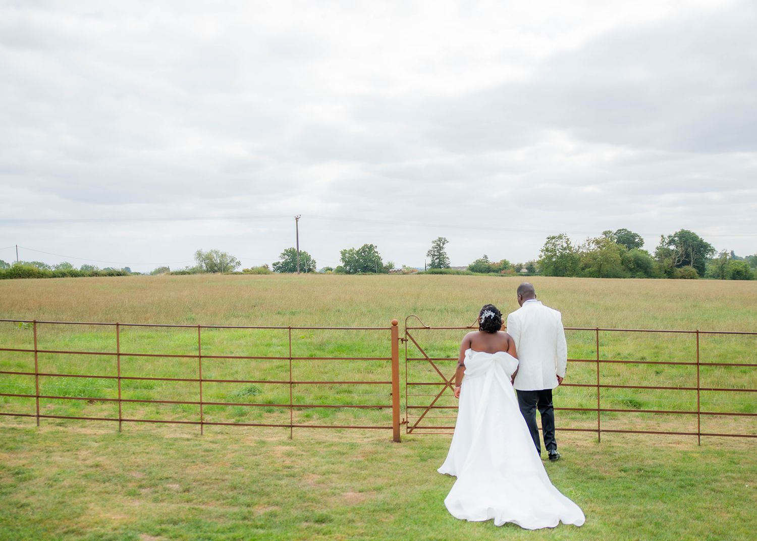 A romantic wedding couple standing by a rustic farm gate in a countryside setting.