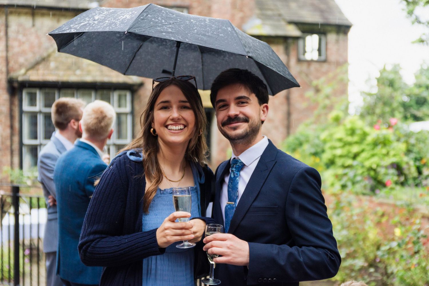 Wedding guests sharing umbrellas and smiling during a light rain at an outdoor celebration.