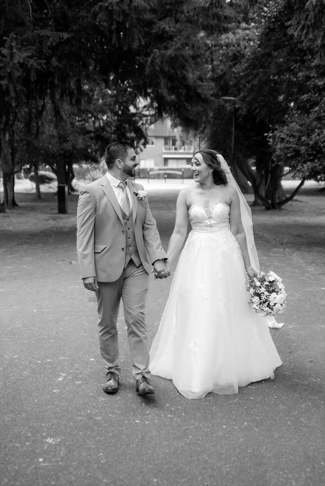 A romantic black and white photo of newlyweds walking hand in hand through a tree-lined path.
