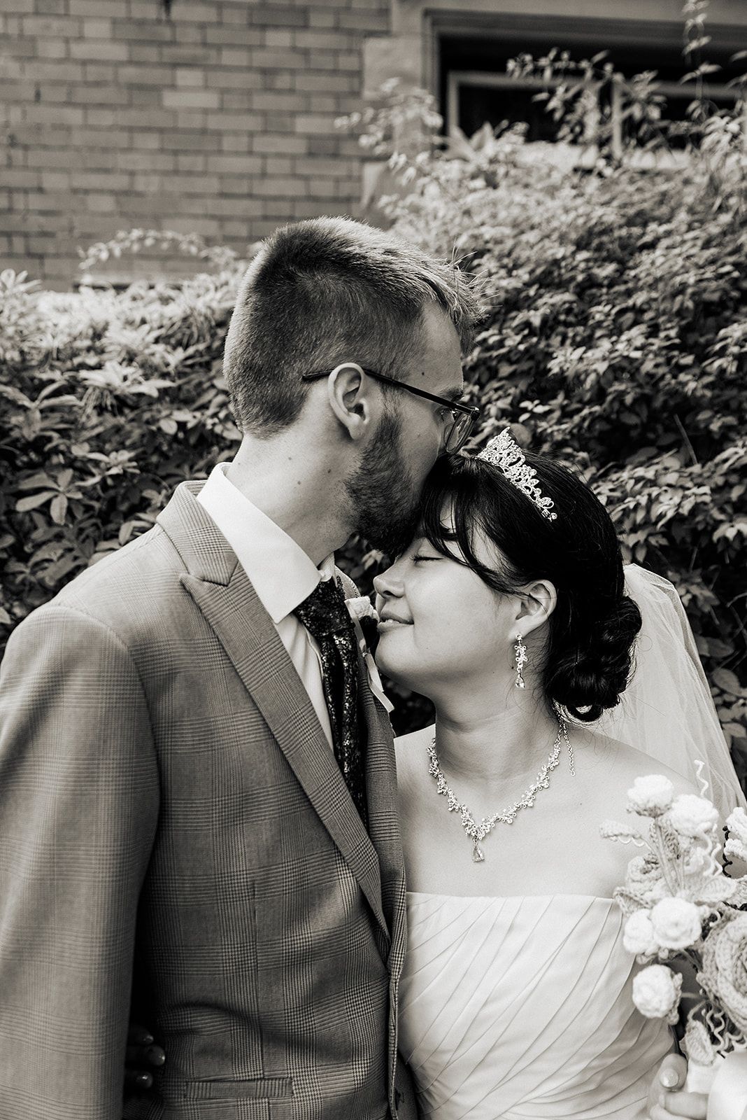 An intimate black and white wedding portrait captured against a brick wall and garden foliage.