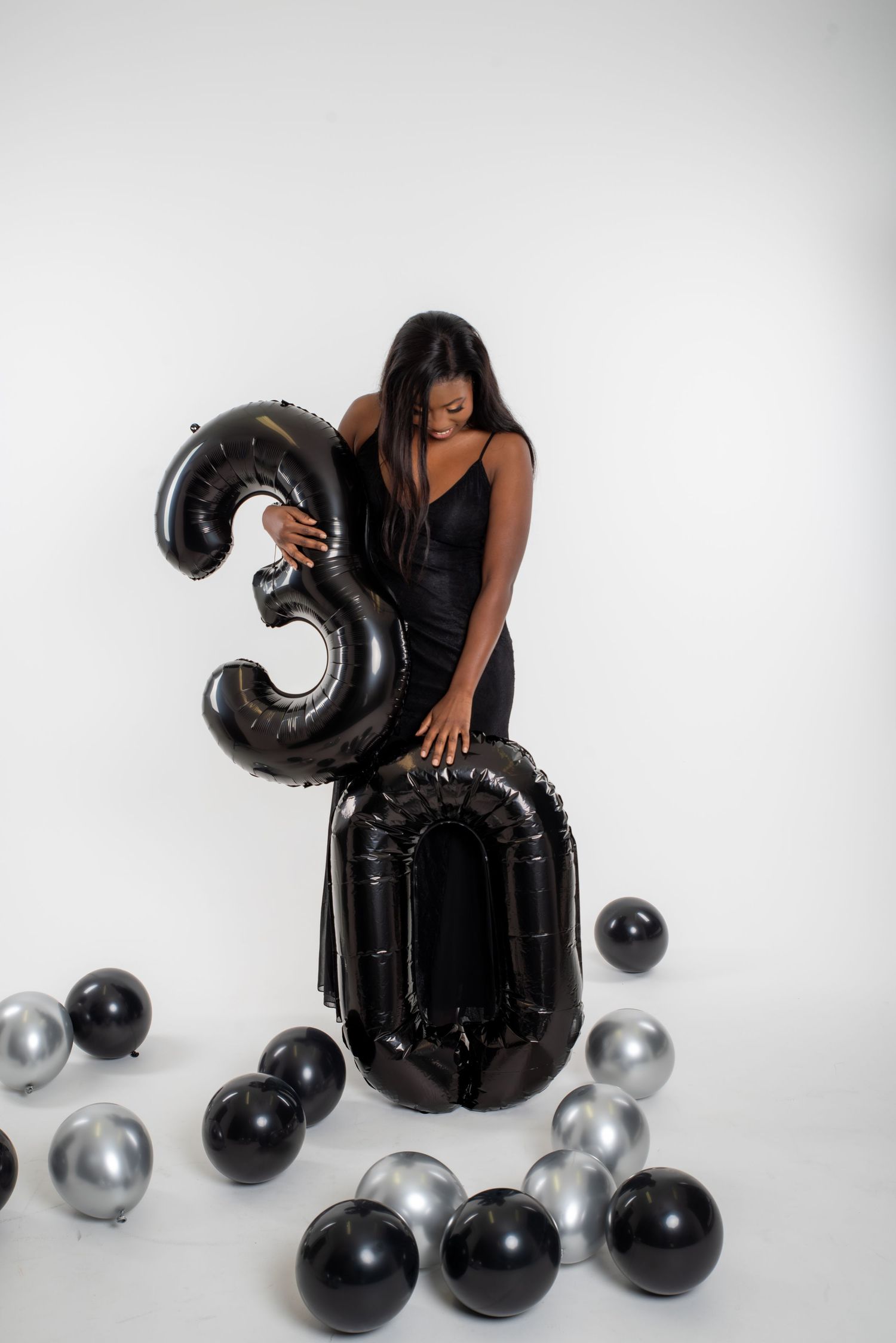 A woman in a black dress poses with black and silver balloons against a white backdrop in a photo series.
