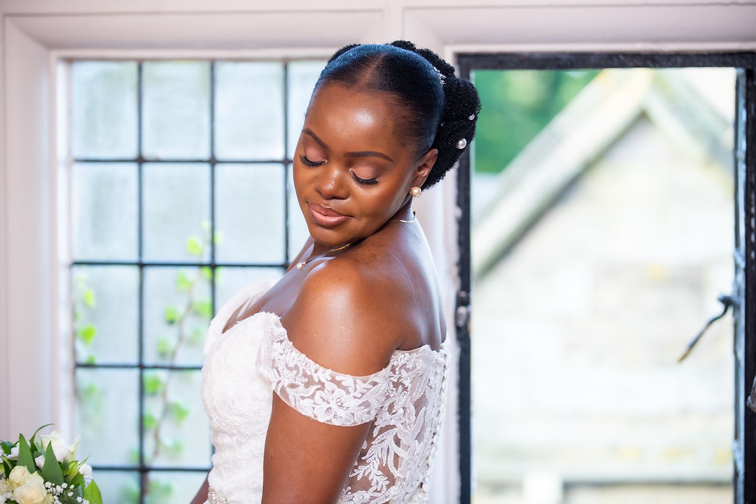 A bride in a white lace gown poses near windows in an elegant indoor setting.