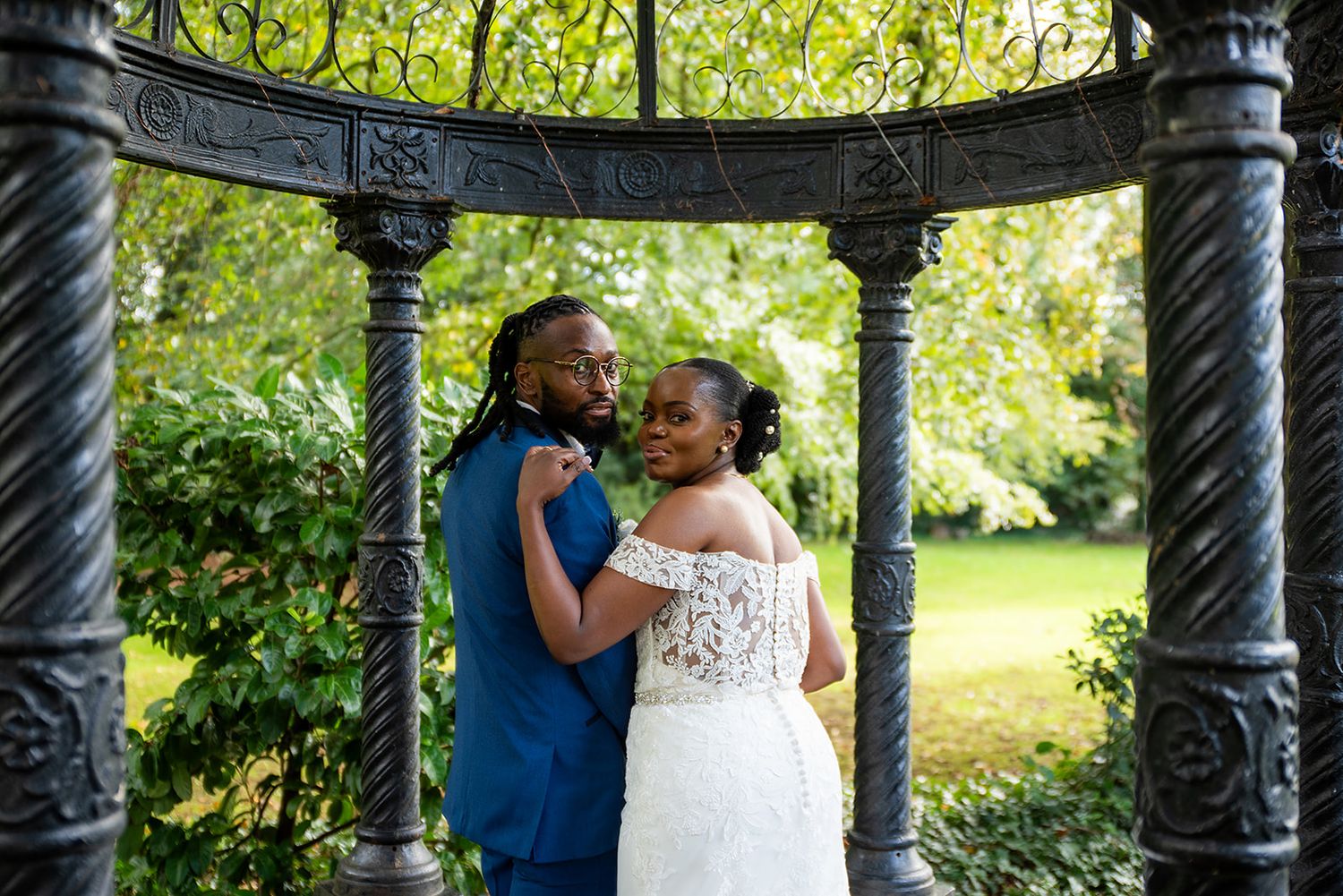 A tender wedding moment captured under an ornate black gazebo in a lush garden setting.