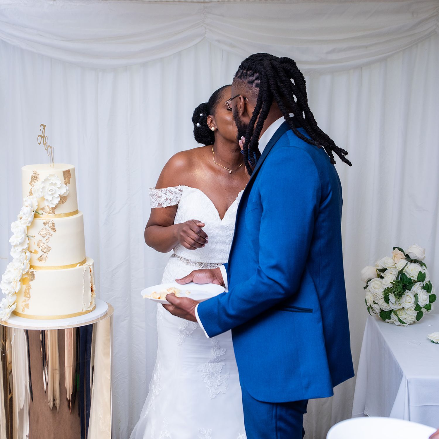 A romantic wedding moment next to an elegant gold-detailed white cake against a draped white backdrop.