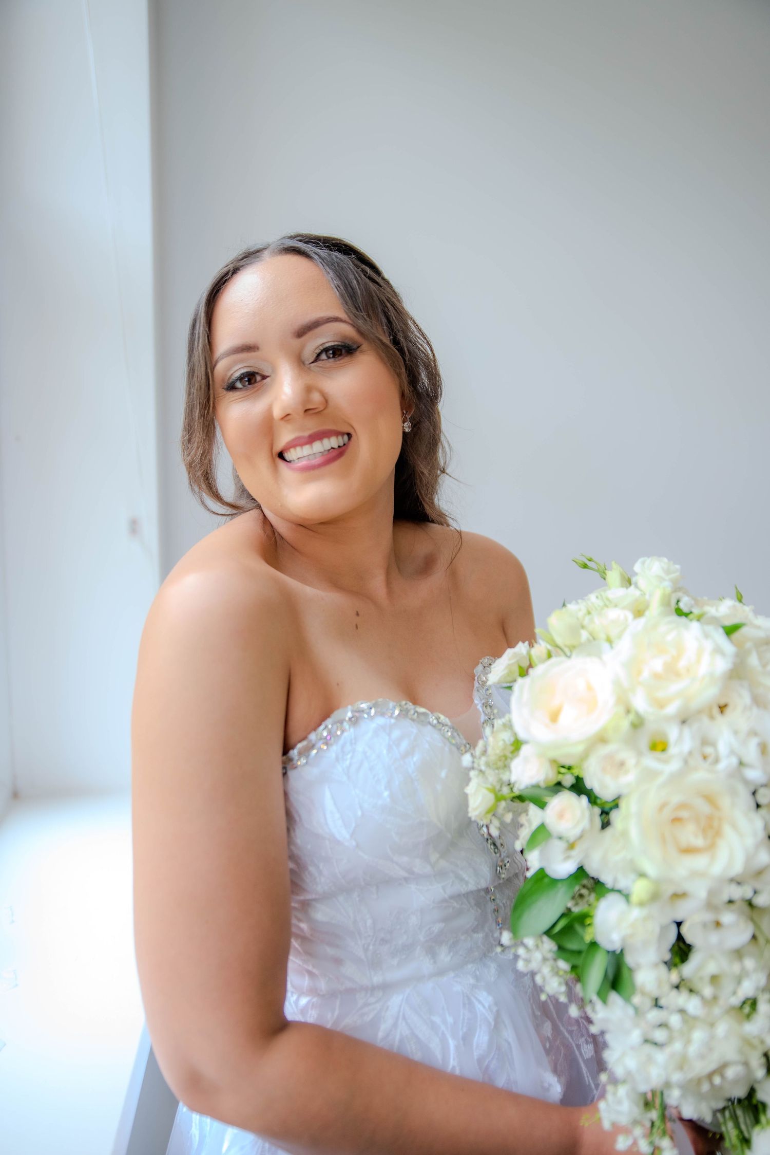 A bride in a white lace dress holds a bouquet of white flowers against a light background.