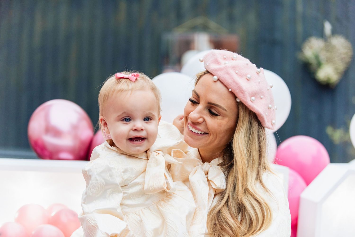 Baby in white dress sharing joyful moment with parent surrounded by pink balloons.