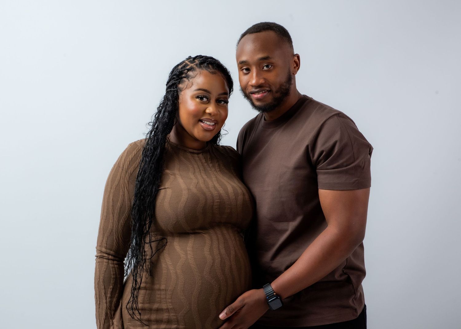 A couple wearing brown outfits poses together for a series of intimate studio portraits against a light gray backdrop.