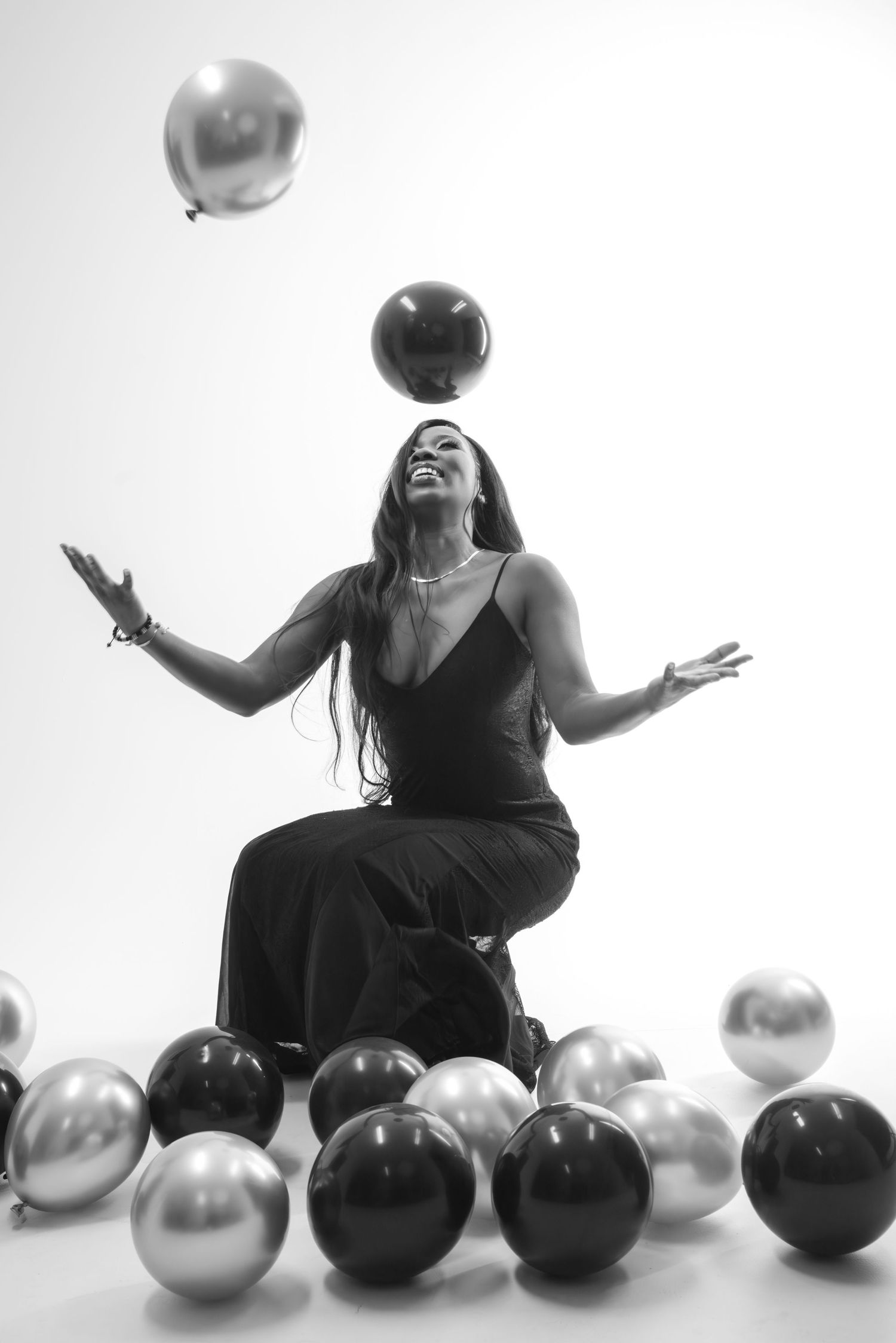 A person in a black dress poses playfully with silver and black balloons against a white background.