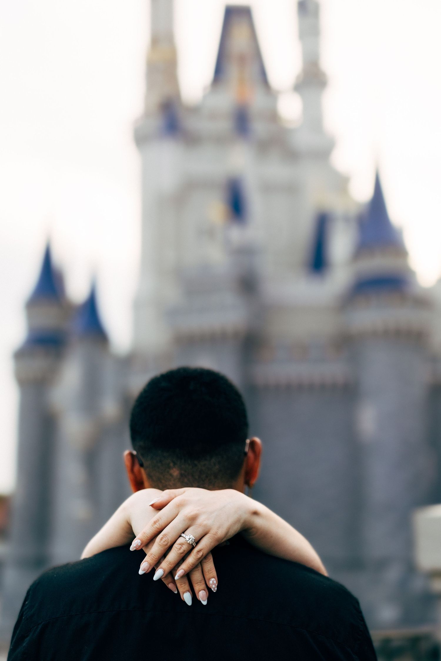 Couple embracing in front of Cinderella Castle at Disney World, woman showing engagement ring on man's shoulder.