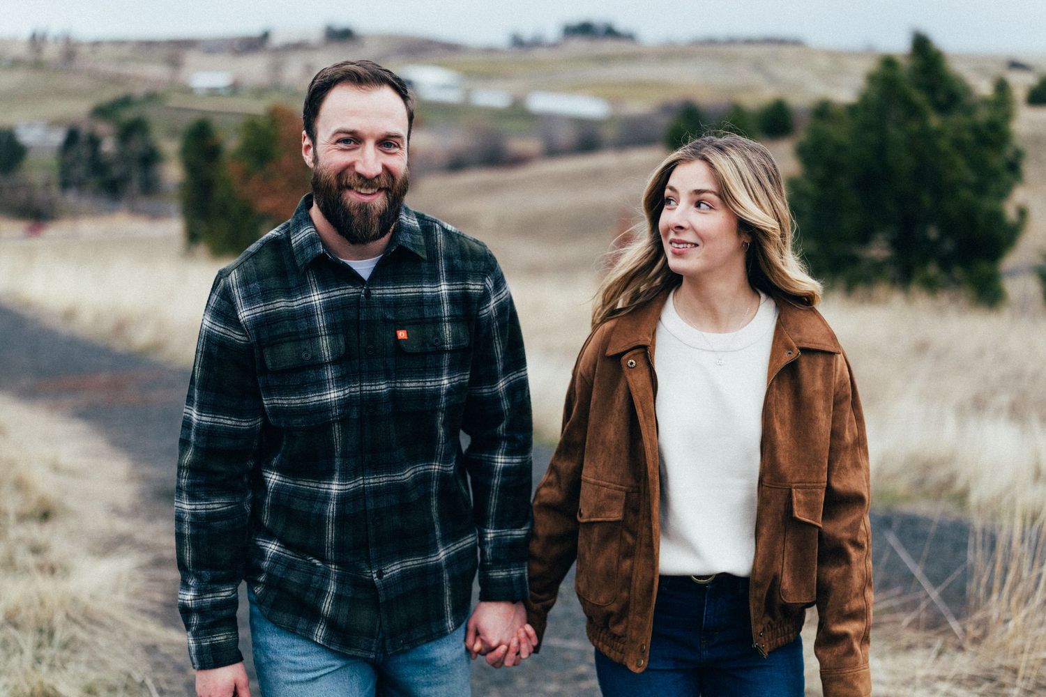 A couple walking hand in hand through a scenic field wearing casual fall attire.