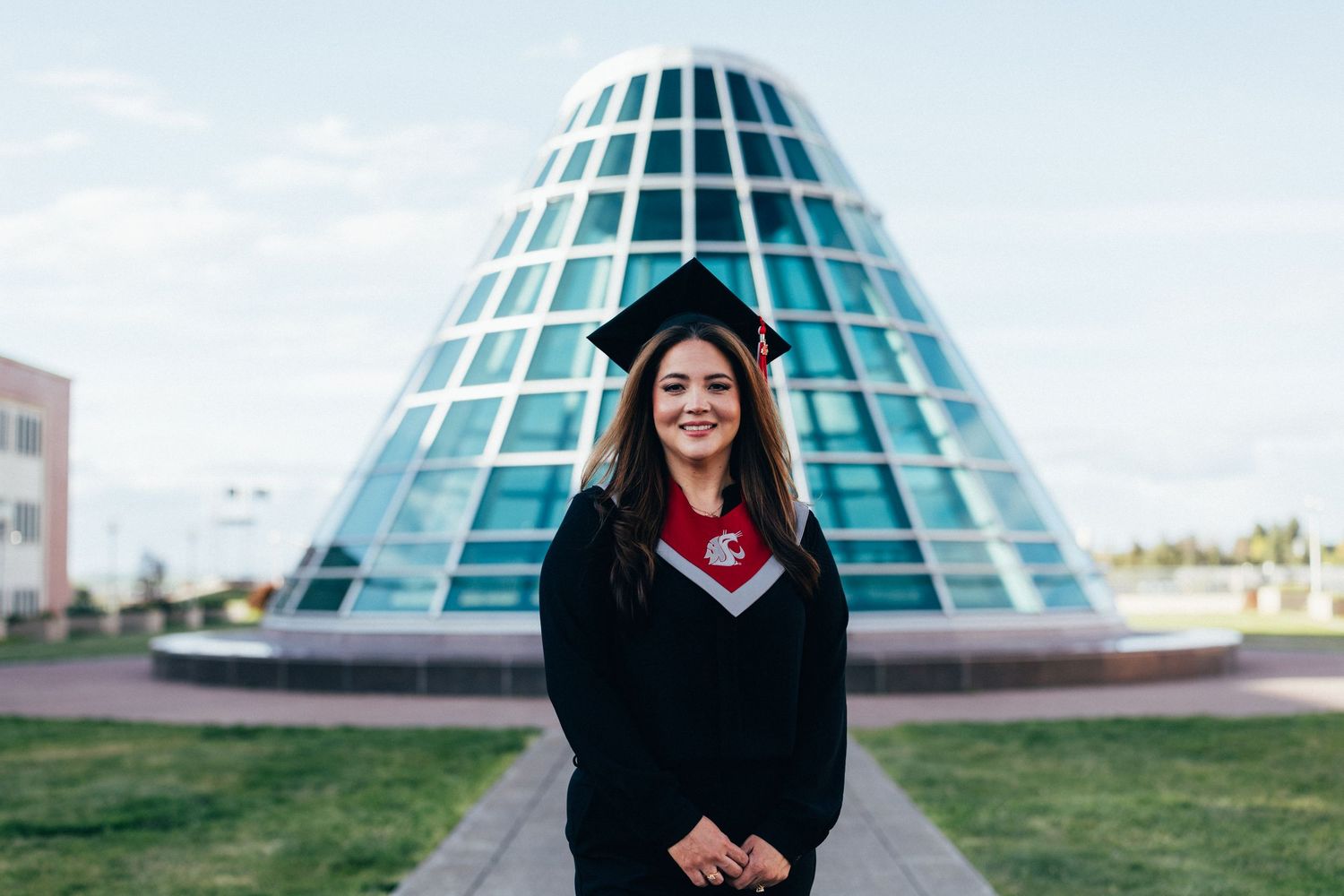 Woman in graduation cap and gown smiling in front of a modern glass pyramid building on a sunny day.