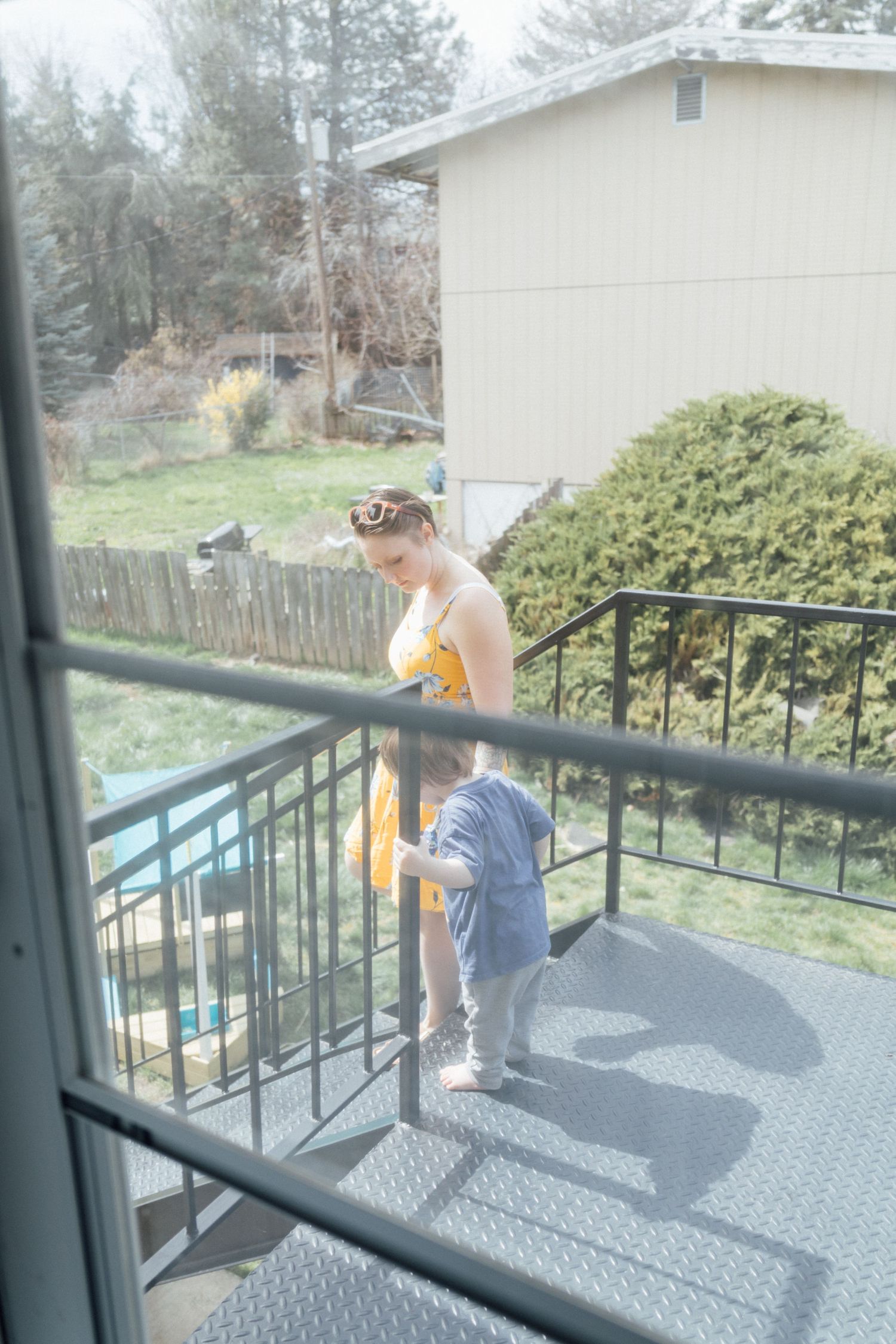 Woman in yellow dress helping toddler walk on outdoor metal staircase with backyard and house in background.