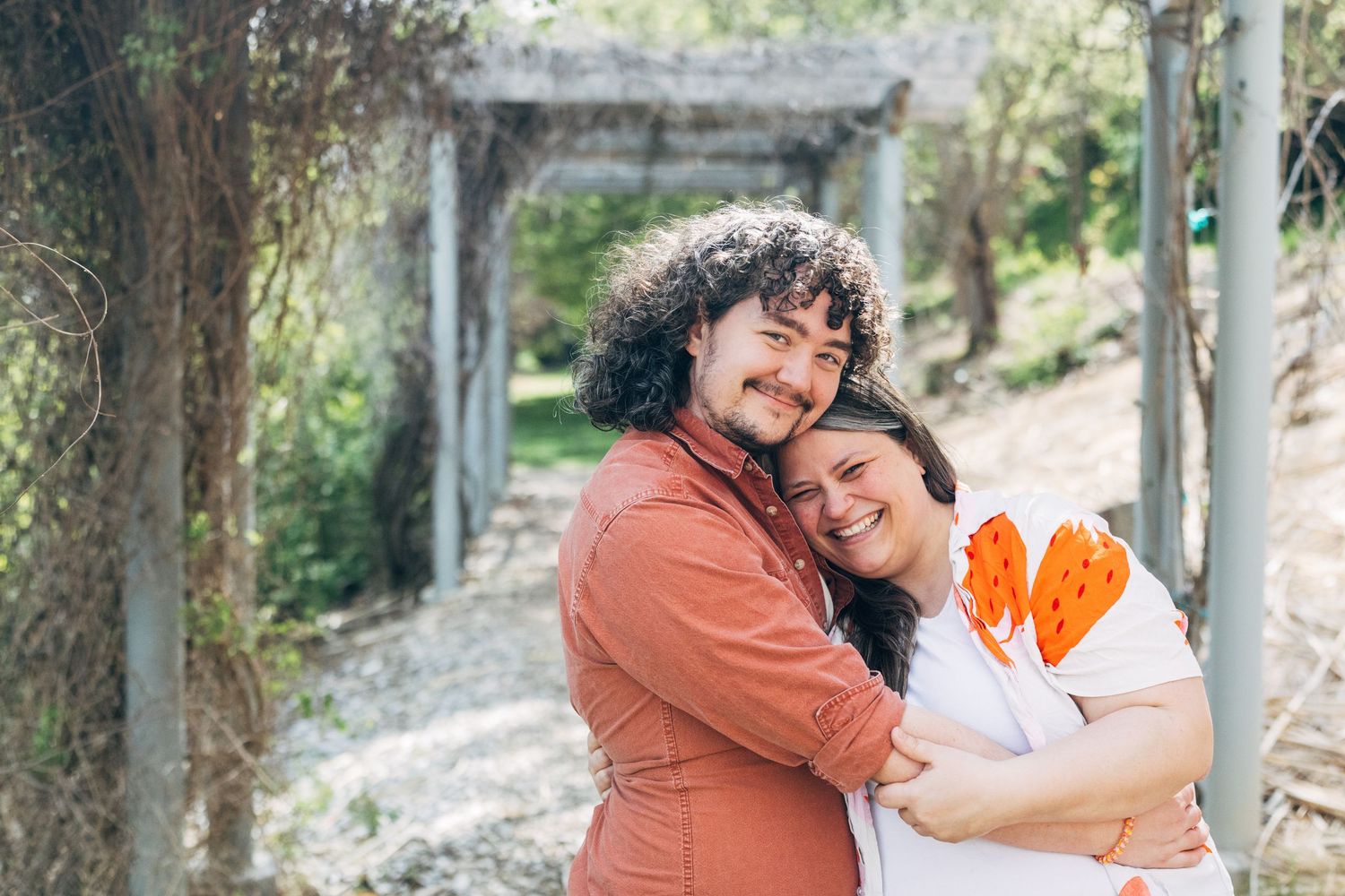 A happy couple embraces outdoors, man in red shirt hugging smiling woman in white tee with orange print, greenery behind.