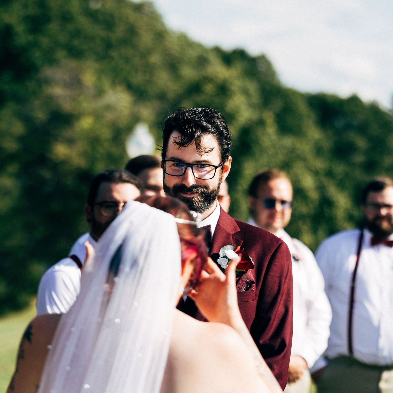 Groom in burgundy suit smiling at bride during outdoor wedding ceremony with groomsmen in background.