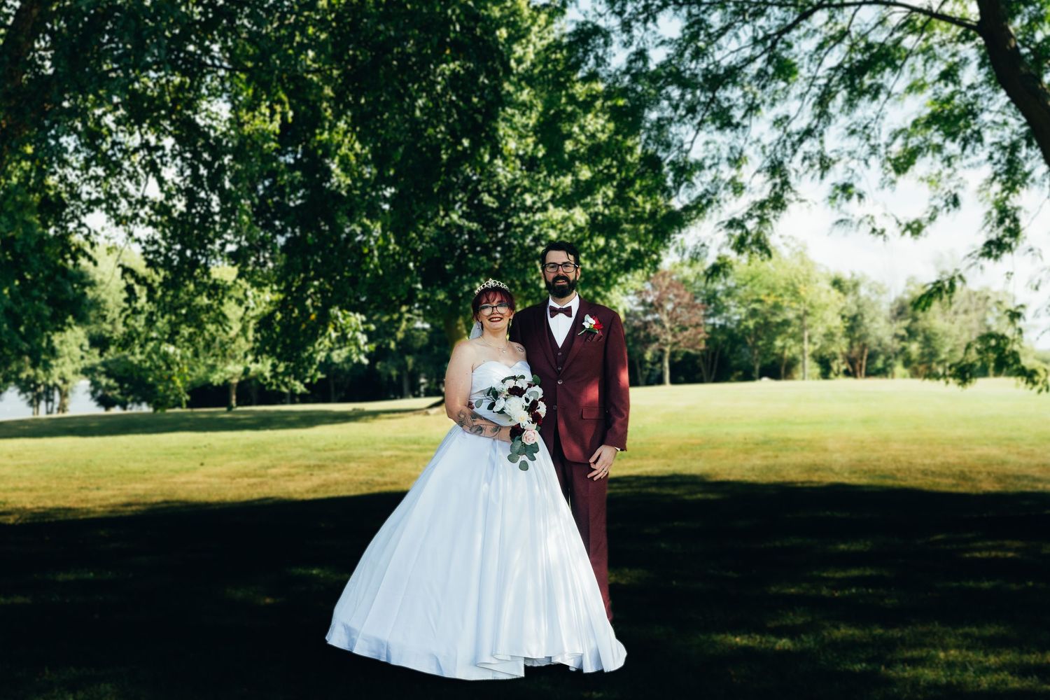 Bride in white ball gown and groom in burgundy suit pose together under lush green trees in a sunlit park.