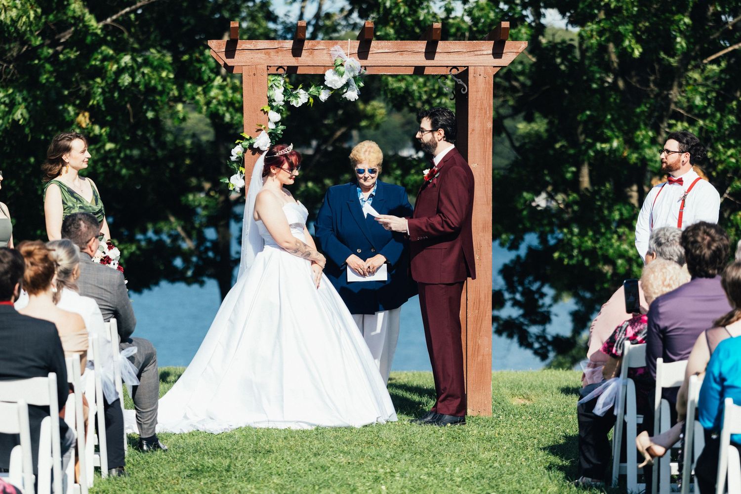 Bride and groom exchange vows under wooden arch outdoors near lake with wedding guests seated on white chairs.