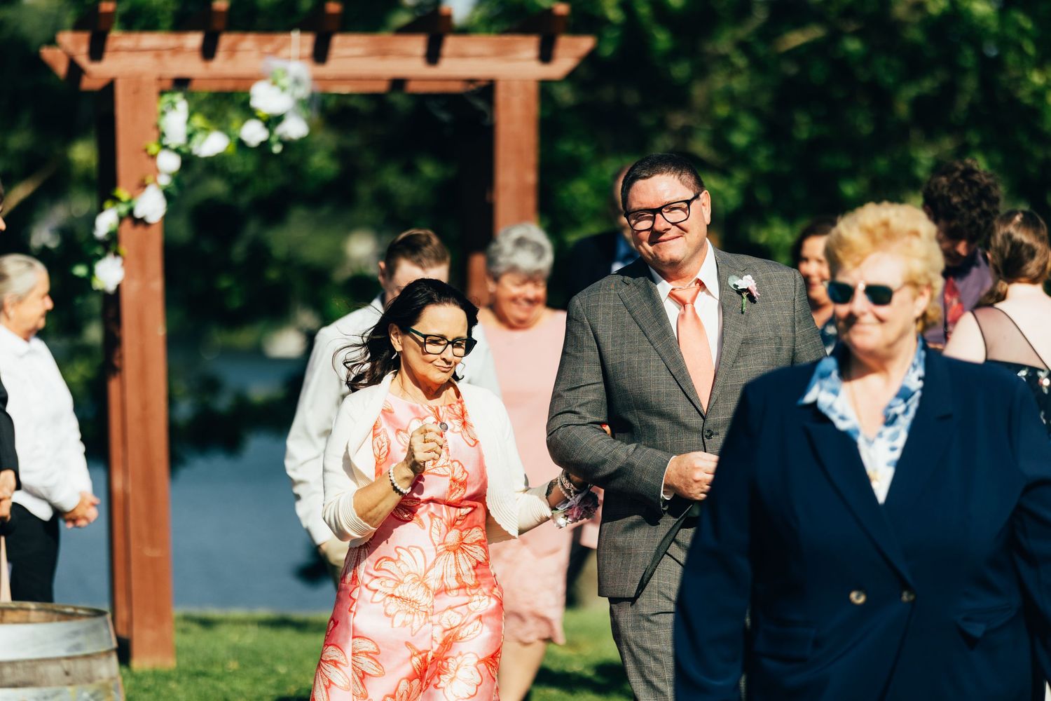 Guests smile and walk at an outdoor wedding ceremony with a wooden arbor backdrop on a sunny day.