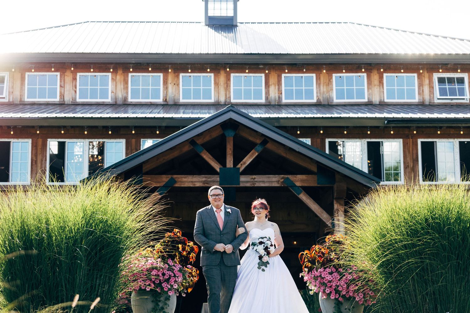 Bride and groom stand outside a rustic barn wedding venue surrounded by fall flowers and ornamental grasses.