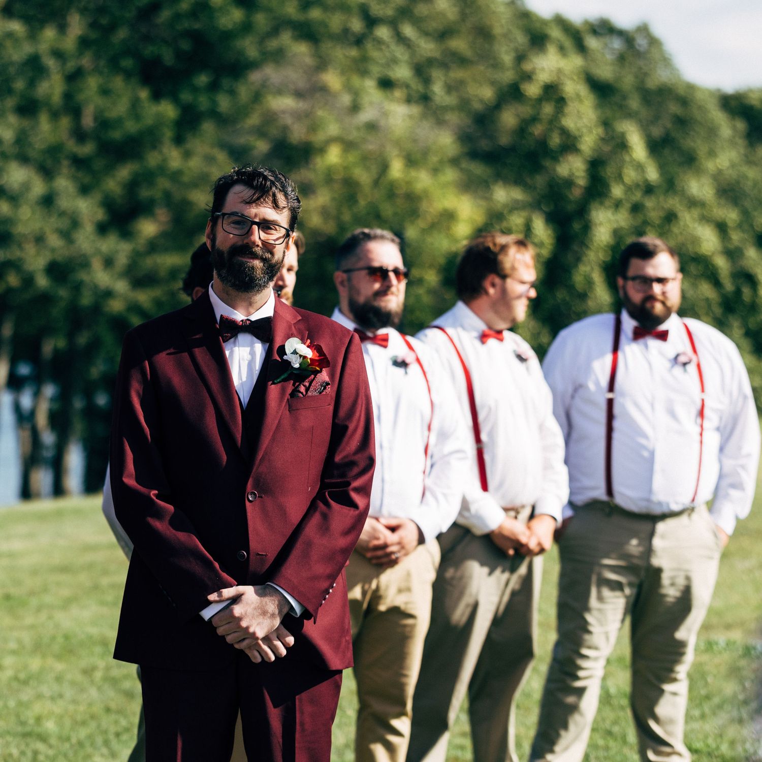 Groom in burgundy suit with groomsmen in white shirts and red bow ties at outdoor wedding ceremony.