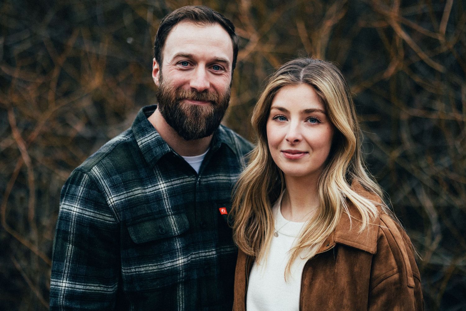 A couple poses together outdoors wearing coordinating casual fall attire.
