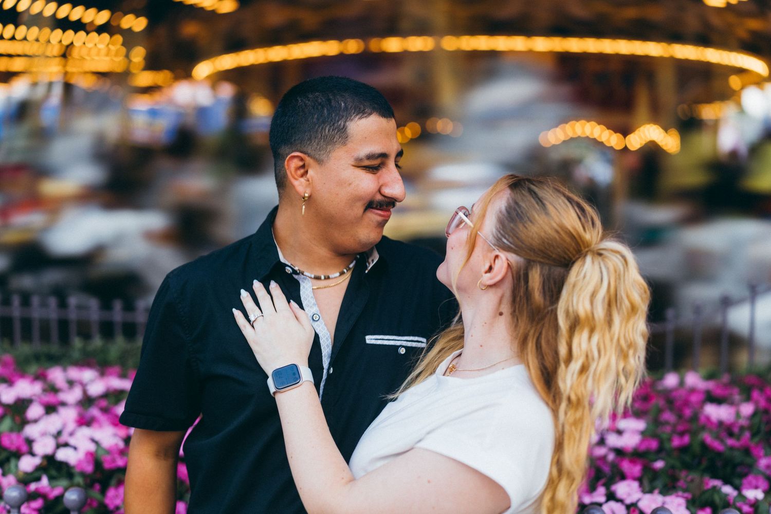 Couple sharing a romantic moment at a carousel fairground, surrounded by colorful flowers and bokeh lights.