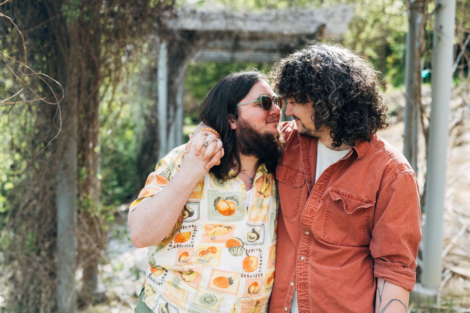 Two curly-haired people in colorful shirts share a joyful moment outdoors, smiling at each other in a garden setting.