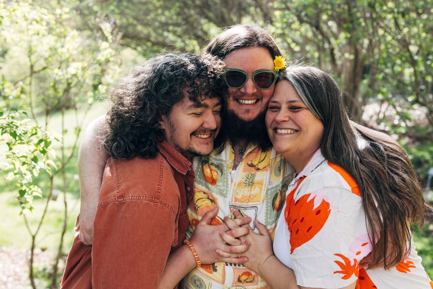 Three friends hugging and smiling outdoors in a lush green park setting on a sunny day.