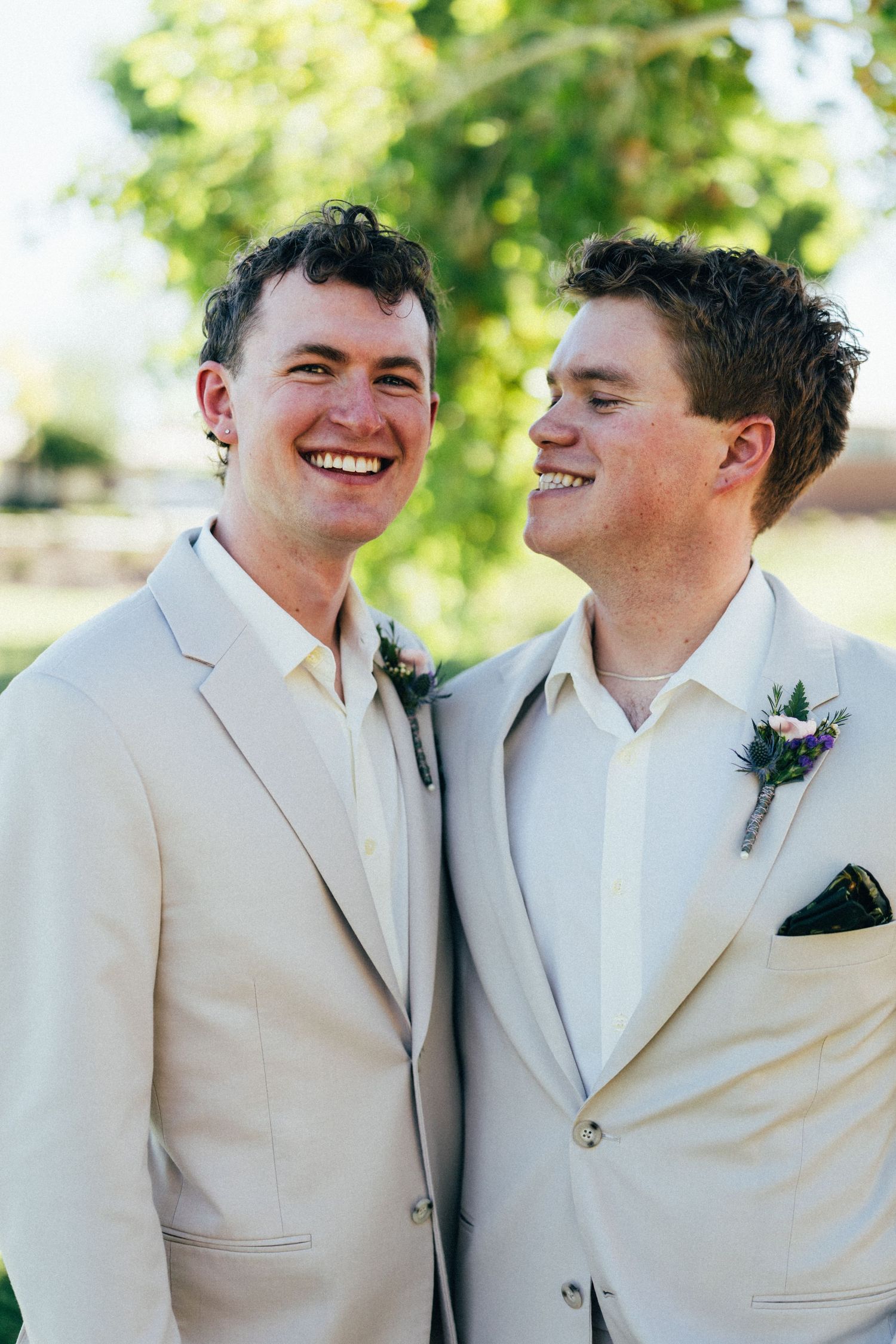 Two grooms in matching cream suits with boutonnieres smile joyfully at each other outdoors on their wedding day.