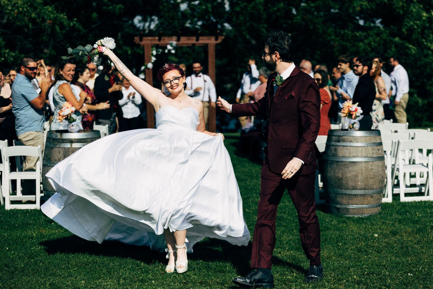 Joyful bride in white dress and groom in burgundy suit dancing outdoors at rustic vineyard wedding celebration.