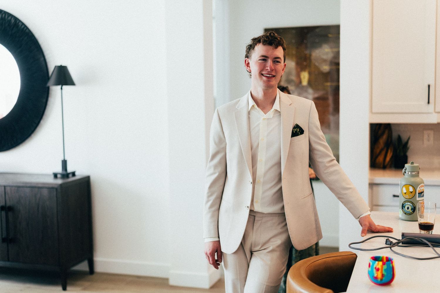 Smiling young man in a beige suit leaning against a kitchen counter indoors.