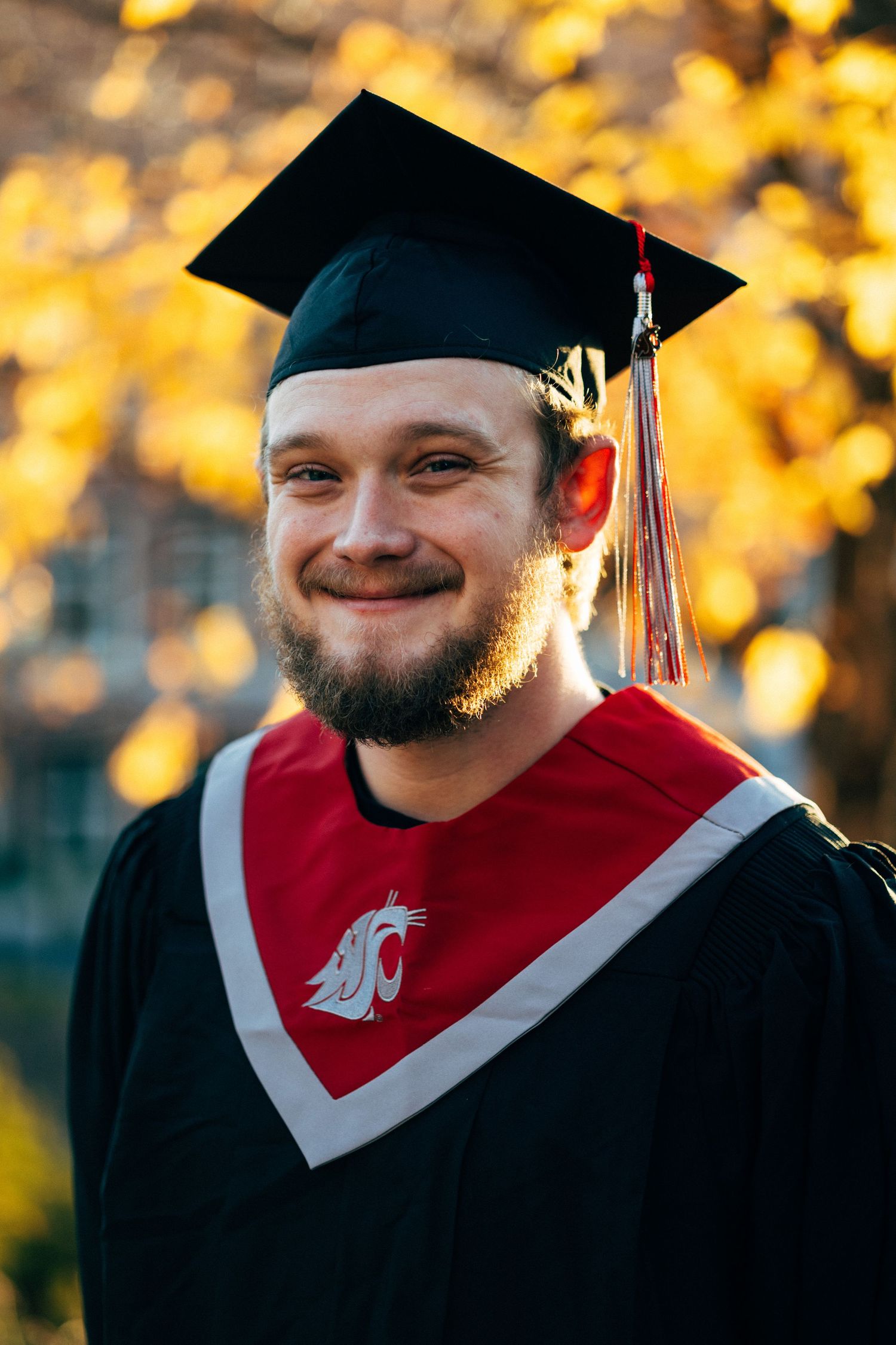 Graduate in red and black academic regalia smiling against autumn foliage background.