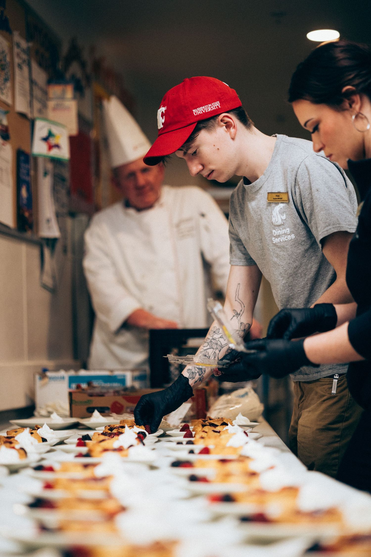 Kitchen staff plating desserts with whipped cream and berries while a chef in white uniform supervises in the background.