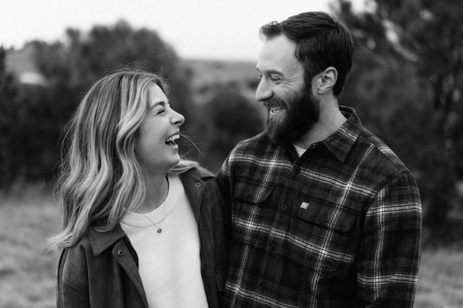 Black and white photo of a smiling couple laughing together outdoors in a field, woman with blonde hair, man with beard.