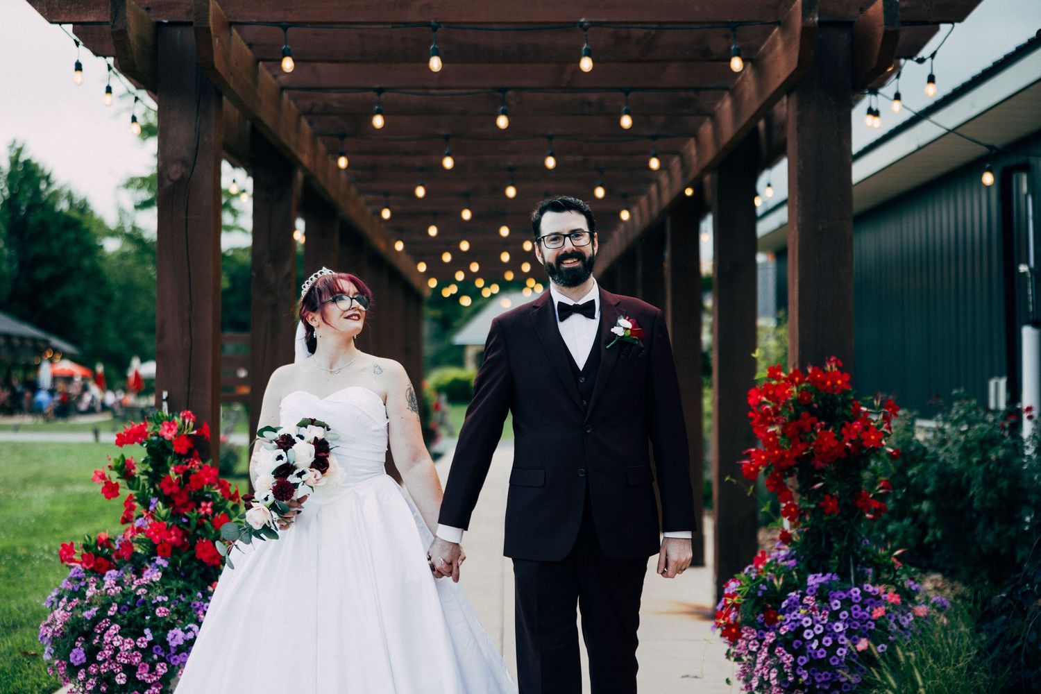 Bride and groom holding hands walking under string lights with colorful floral arrangements at outdoor wedding venue.