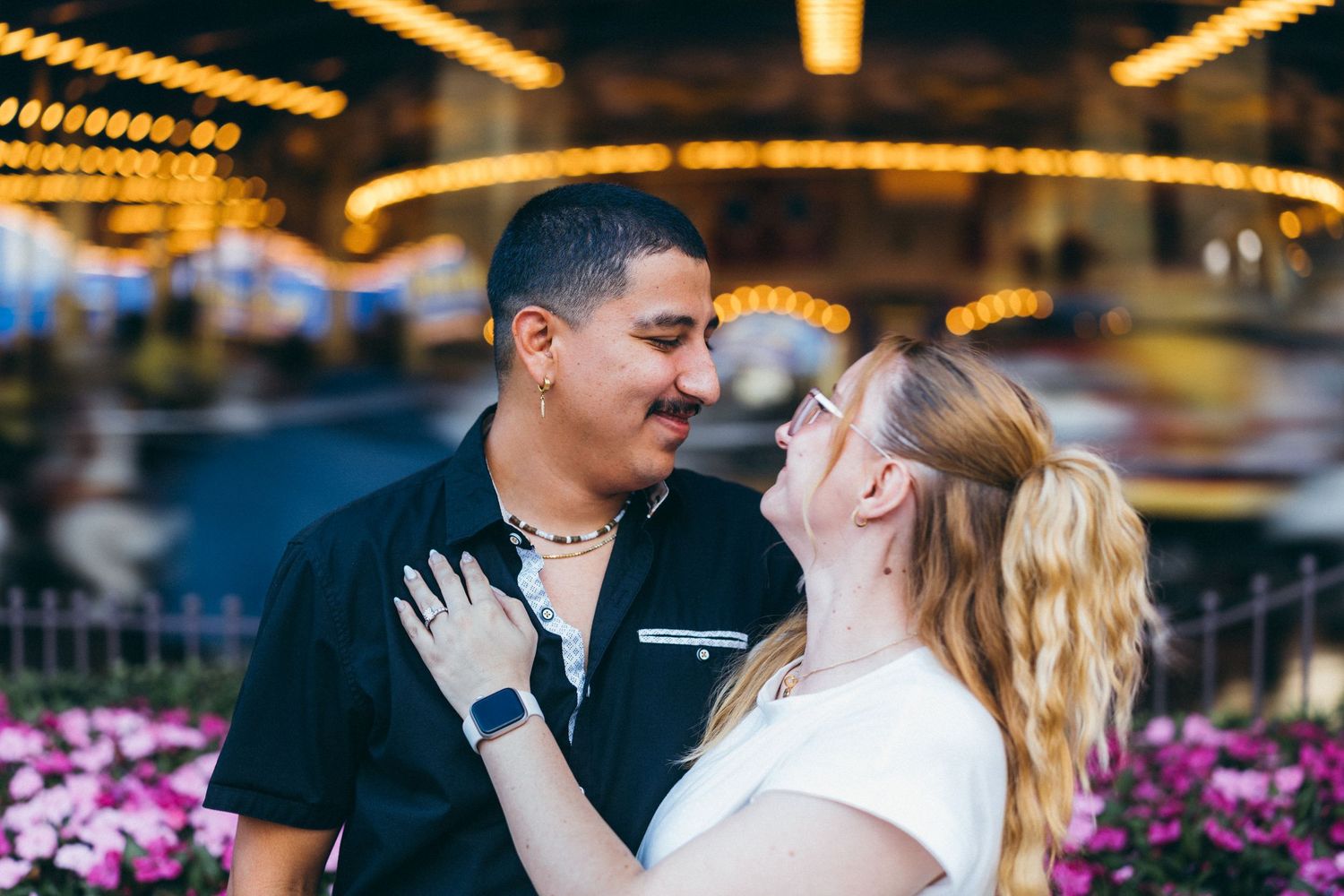 Couple sharing a romantic moment in front of a glowing carousel at an amusement park at night.