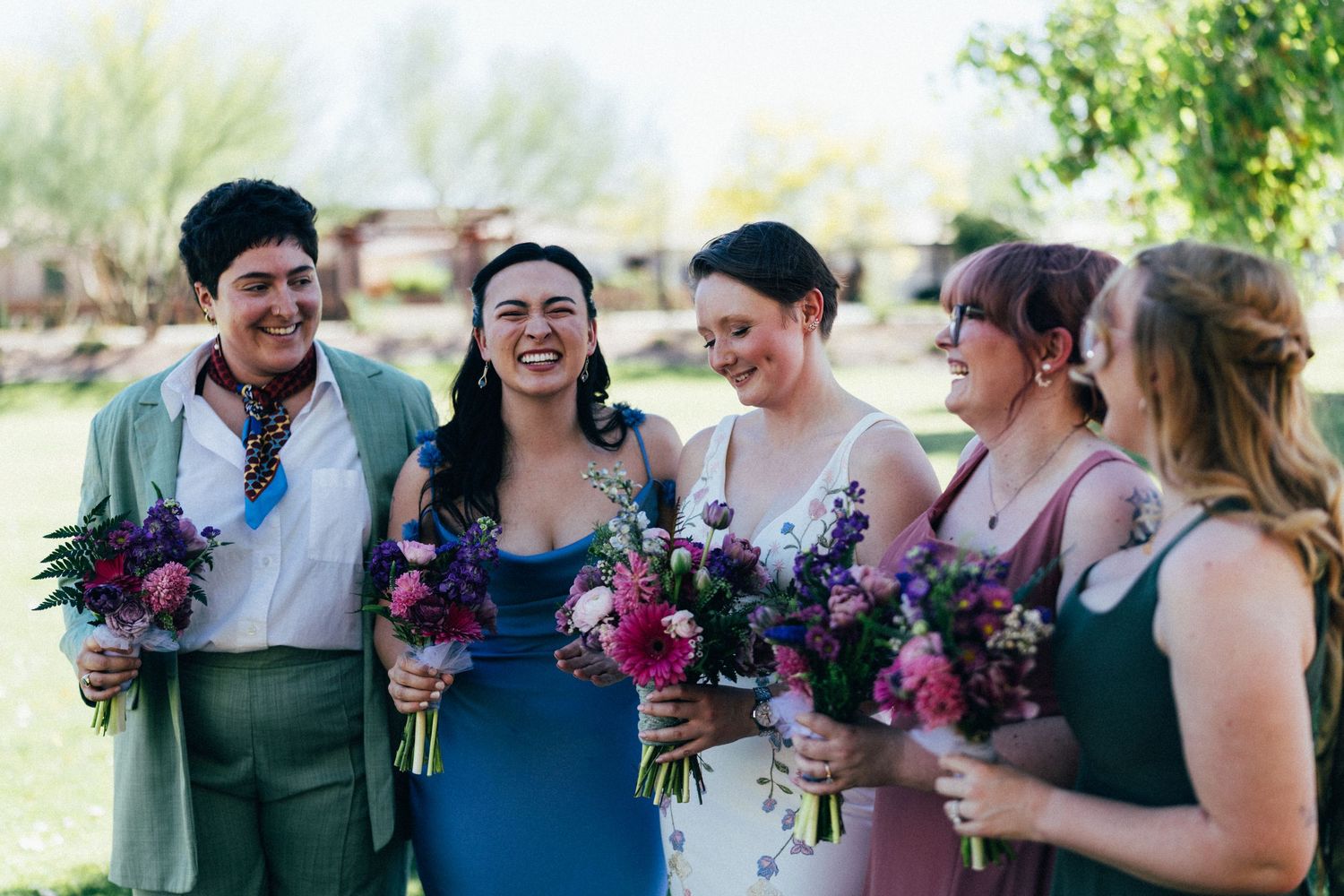 Bridal party laughing outdoors, holding colorful bouquets in blue, green, and mauve dresses.